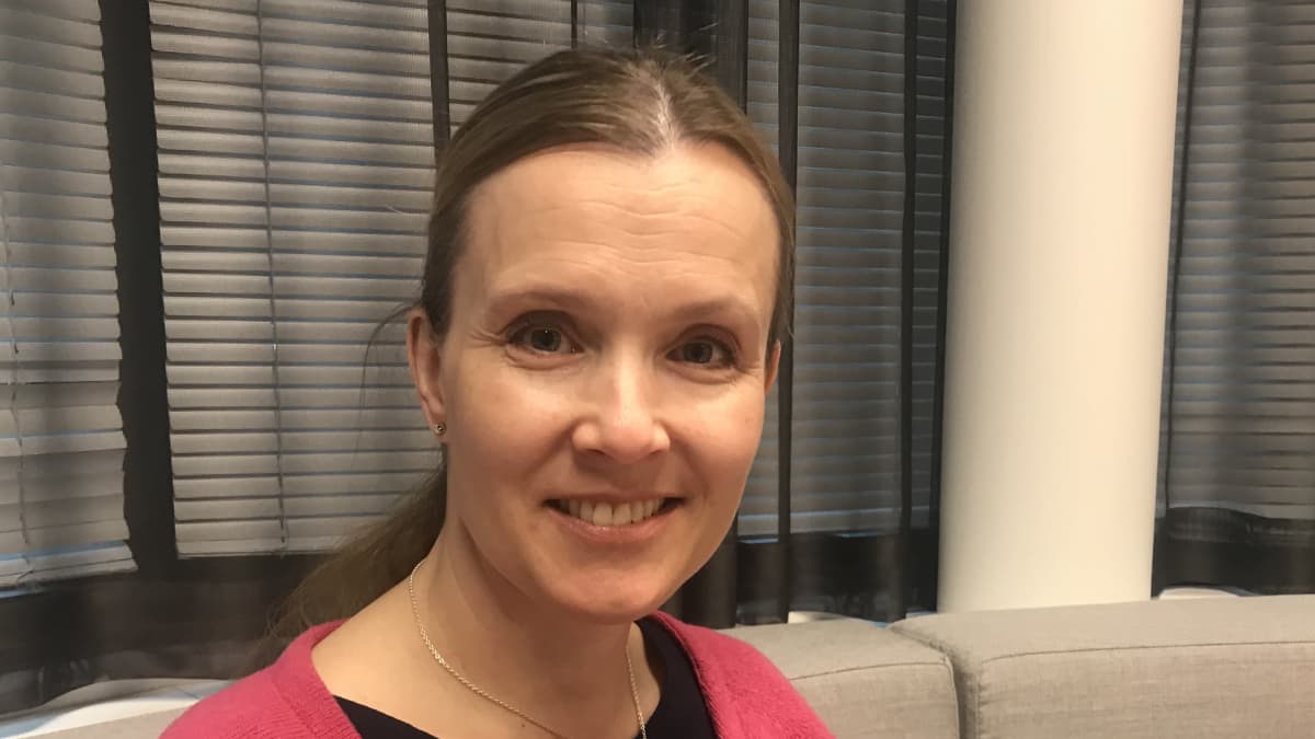 Nainen istuu harmaassa sohvassa ja hymyilee. Naisella on musta paita ja sen päällä punainen neuletakki.