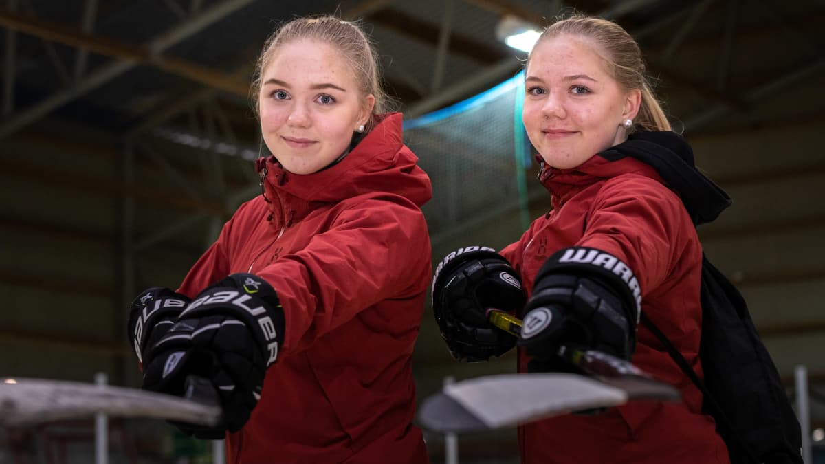 Iida (vas.) ja Emma Lappalainen punaisissa takeissa, jääkiekkohanskat kädessä, ojentamassa jääkiekkomailoja kameraa kohti.