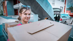 Juuli Kyllönen kantaa pahvilaatikkoa ja katsoo kameraan. Taustalla auto ja peräkärry.