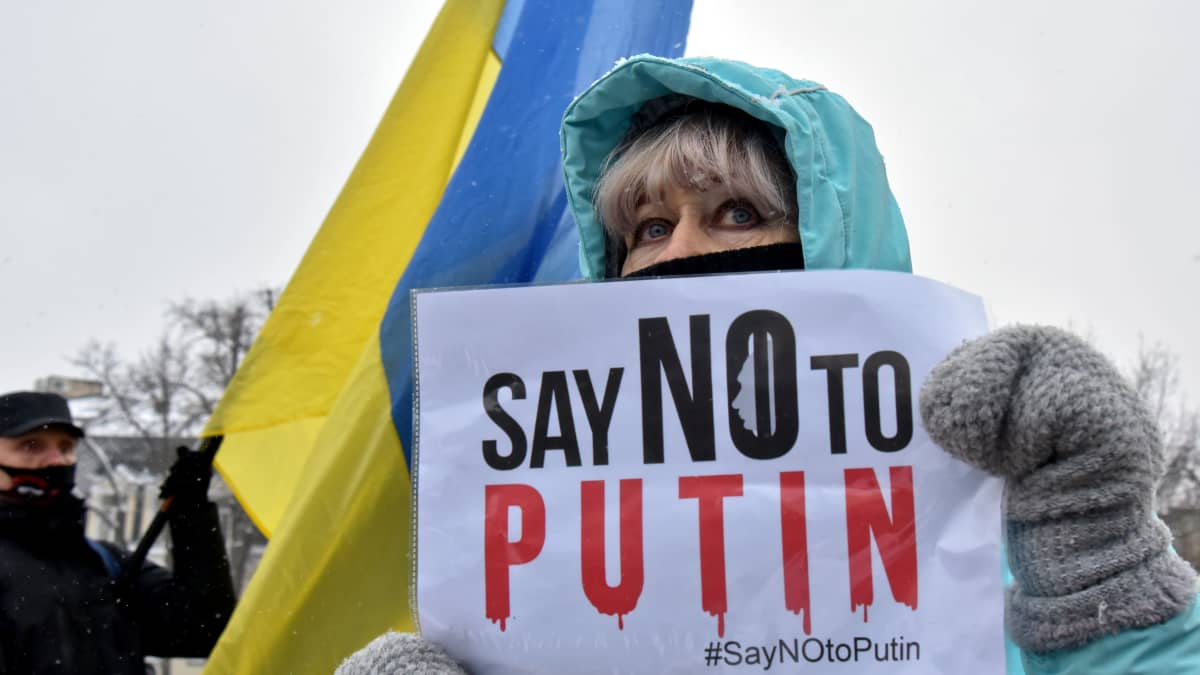 Nainen pitää käsissään kylttiä, jossa lukee "Say no to Putin". Taustalla näkyy keltasininen Ukrainan lippu.