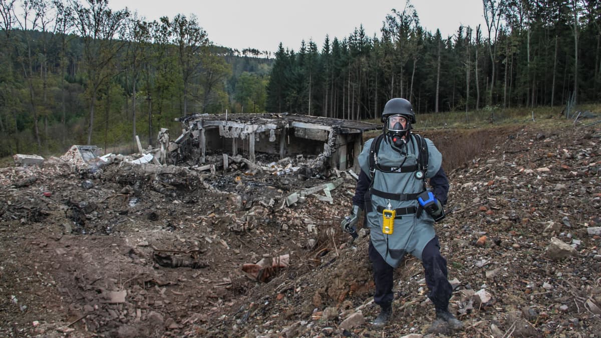 Suojapukuun, kaasunaamariin ja kypärään pukeutunut mies seisoo räjähtäneen näköisessä maisemassa tuhoutuneen betonisen rakennelman edessä.Taustalla näkyy metsämaisemaa.