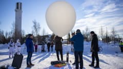 Joukko miehiä virittelee isoa valkoista ilmapalloa satelliitin kannattimeksi aurinkoisessa talvisäässä, runsaasti lunta ja Seinäjoen Lakeuden Risti näkyy taustalla.