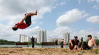 Nuori mies suorittaa akrobaattista voltin ilmaan hiekkarannalla, taustalla korkeita rakennuksia ja muita ihmisiä nauttimassa aurinkoisesta päivästä.