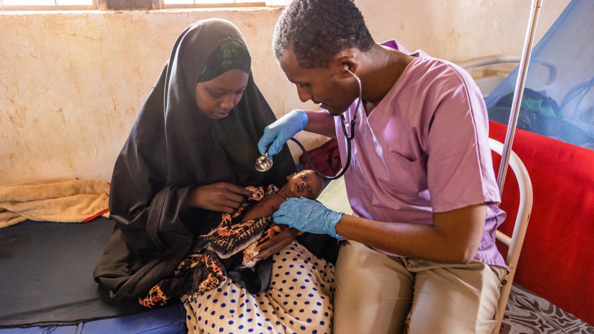 Lääkäri tutkii mustakaapuisen nuoren äidin sylissä olevaa laihaa vauvaa stetoskoopilla somalialaisella aliravitsemusklinikalla
