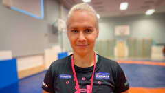 Vaaleahiuksinen, mustaan t-paitaan pukeutunut Petra Olli seisoo urheilusalissa kädet selkänsä takana ja hymyilee kameralle.