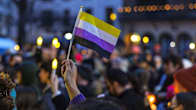 A person's hand holding up a small yellow, white, purple and black-striped flag, with a large crowd of people seen, out of focus, in the background.