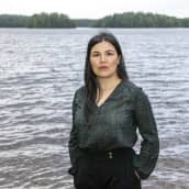Mustatukkainen nainen seisoo koivun vieressä järven rannalla.