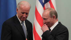 Biden ja Putin seisovat mustissa puvuissa Yhdysvaltain lipun edessä. Putin hymyilee ja pitää kättään leuan ja suunsa edessä. Biden näyttää oikealla kädellään peukaloa.