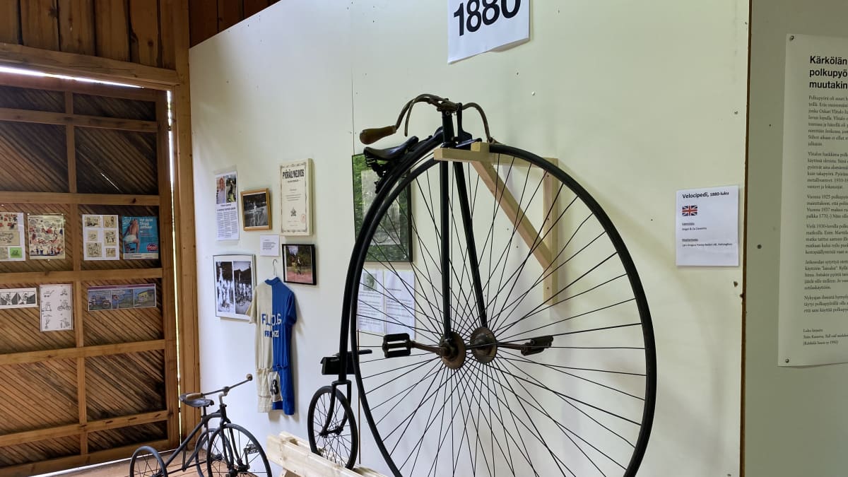 Isolla etupyörällä ja pienellä takapyörällä varustettu vanhanmallinen polkupyörä, jonka takana vastaava pienempi, lasten malli mutta kahdella pienellä takapyörällä. Seinll kehystettyjä kuvia, sinivalkoinen pyöräilypaika ja vuosiluku 18880.