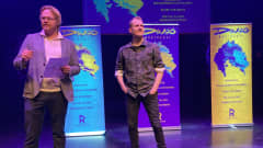 Kolmen Dingo-julisteen edessä kaksi miestä seisoo teatterin lavalla.