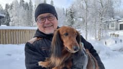Mies koira sylissään talvisessa lumimaisemassa
