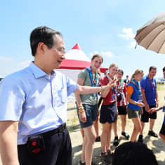 A smiling middle-aged man in glasses and a blue short-sleeved shirt talks to about 7 teenagers, mostly girls wearing shorts, T-shirts and bandanas, one holding a parasol. 