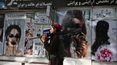 Taliban sotilas kävelee kaupan ohitse missä naisten kasvot on peitetty mustalla maalilla.