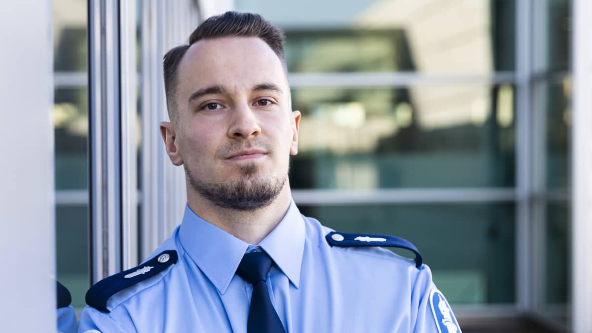 Nuori mies poliisin uniformussa nojaa ulkoseinään ja katsoo kameraan kädet puuskassa. Hän hymyilee pienesti.