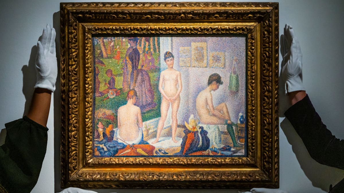 Kaksi käsiparia asettaa maalausta seinälle. Maalauksessa on kolme alastonta naista.