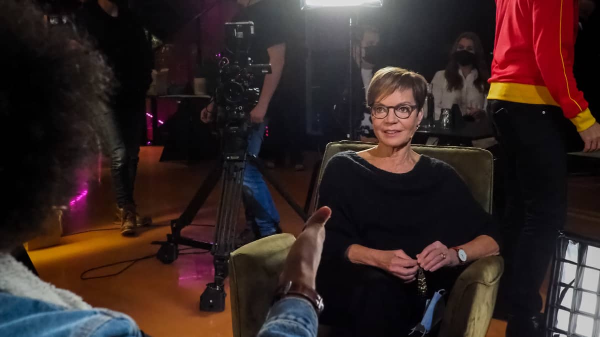 Näyttelijä Lena Meriläinen istuu Tampereen G Livelabin tuoleissa. Perjantain juontaja Sean Ricks keskustelee hänen kanssaan. Taustalla näkyy Perjantai-ohjelman kuvaushenkilöstöä ja -välineistöä.