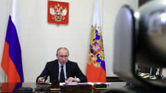Vladimir Putin sitter vid ett mörkt och blankt skrivbord och tittar in i en videokamera.