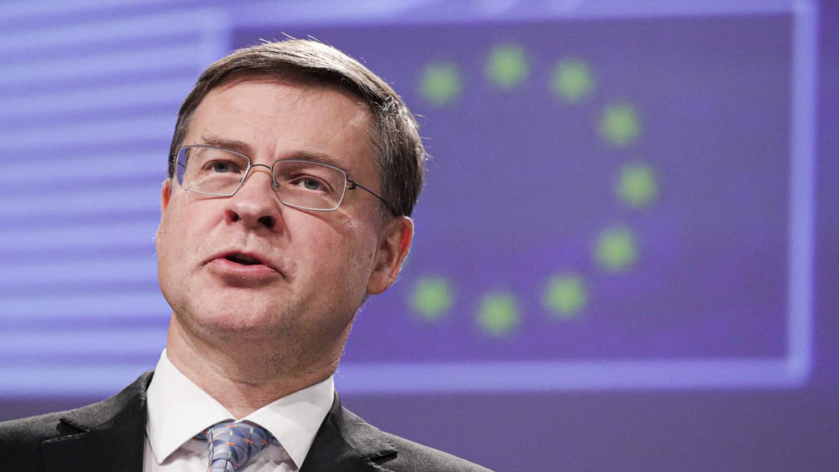 EValdis Dombrovskis, wearing a dark suit and glasses, speaks during a press conference with an EU flag visible dimly in the background.