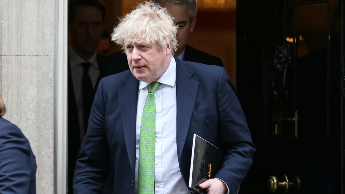 En rufsig blond man i grön slips med en mapp i handen går ut genom en svart dörr.