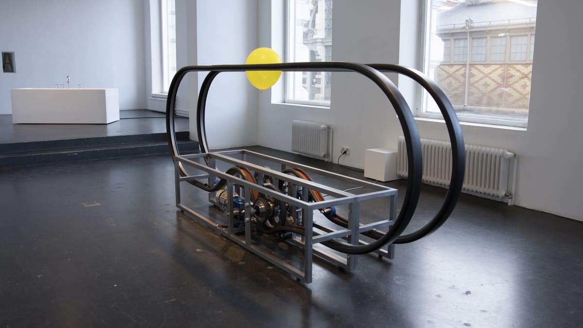 Eetu Huhtalan taideteos on mekaaninen veistos, jossa eri suuntiin pyörivien liukuportaiden kaiteiden väliin jumiin jäänyt keltainen ilmapallo pyörii.