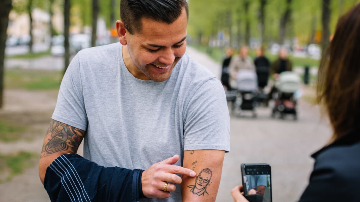 Mies näyttää käsivarrestaan tatuointia kännykkäkameralla kuvaavalle naiselle.