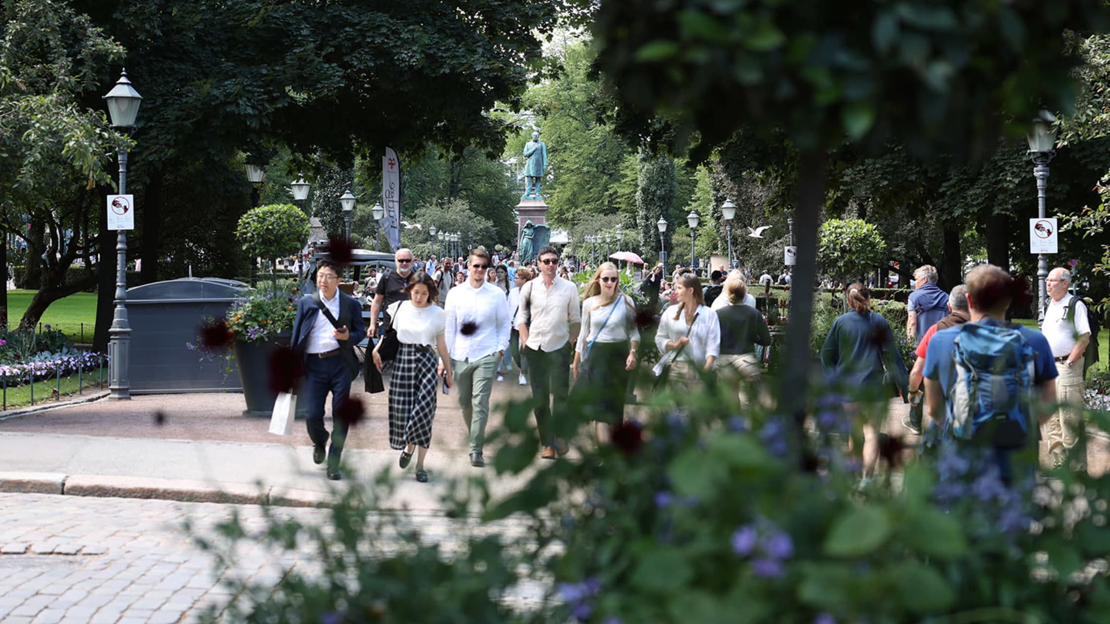 Turisteja Helsingin Esplanadin puistossa kävelemässä kohti kameraa. Etualalla vihreitä kasveja, taustalla patsas.