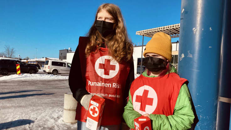 Kaksi lasta pitelee Punaisen Ristin keräyslippaita käsissään.