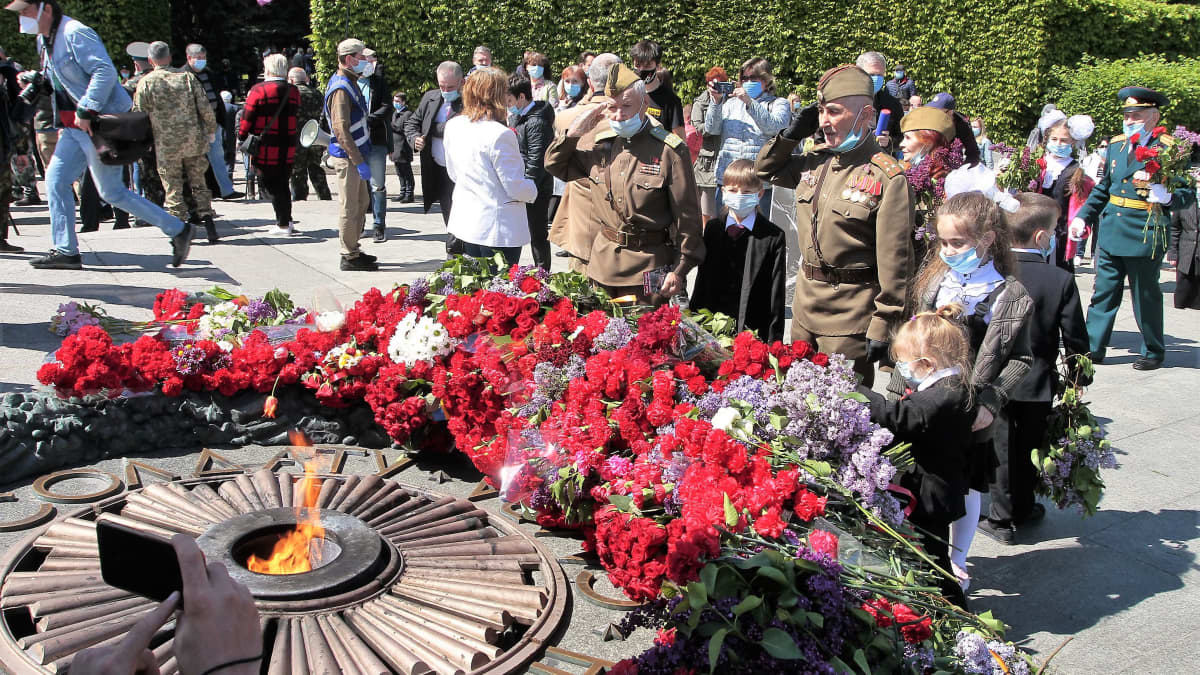 Toisen maailmansodan univormuihin pukeutuneet vanhat miehet tekevät lasten kanssa kunniaa ikuisen tulen äärellä. Muistomerkin juurella on suuri määrä punaisia kukkia.