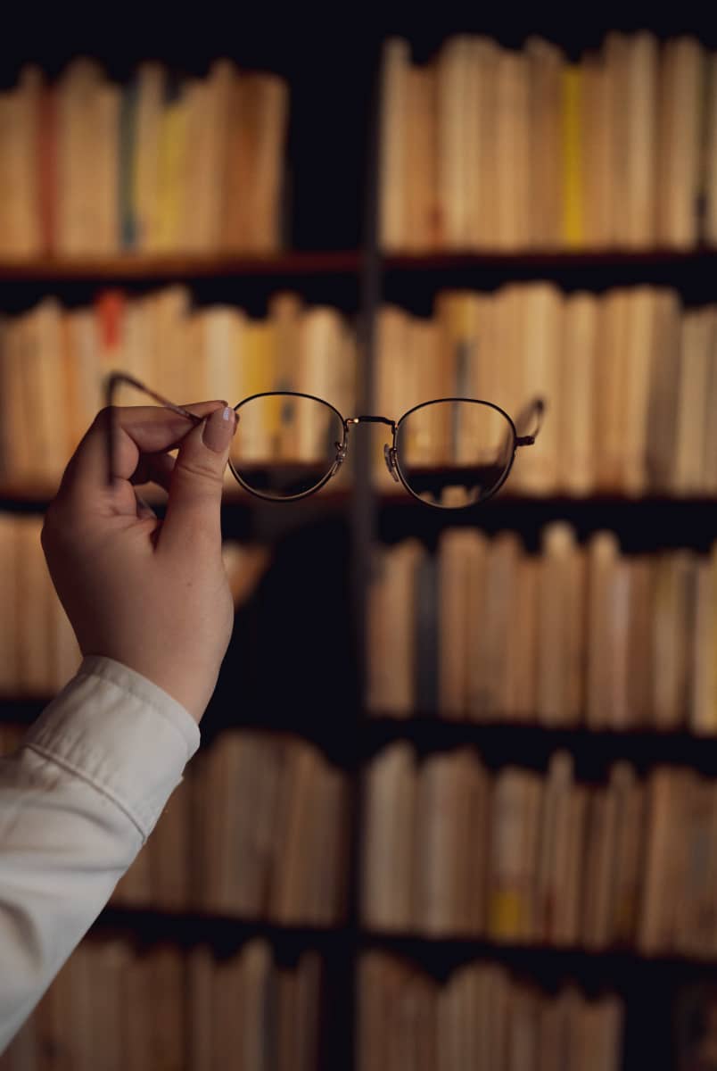 Lähikuva silmälaseista vasten kirjahyllyjä hämärässä kirjastossa.