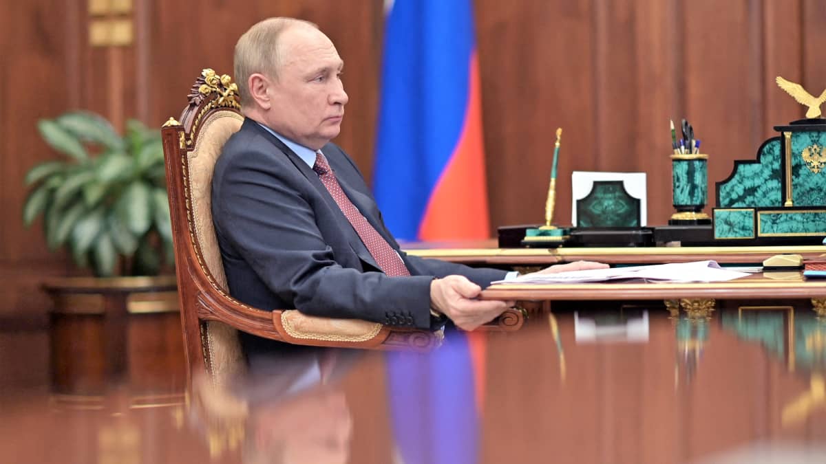 Putin istuu pöydän ääressä tummanharmaassa puvussa ja viininpunaisessa kravatissa. Taustalla huoneessa näkyy Venäjän lippu.
