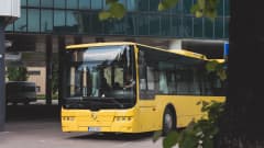 Keltainen bussi kaartaa asemalta kadulle. Kuljettajan vieressä on kaksi matkustajaa. 