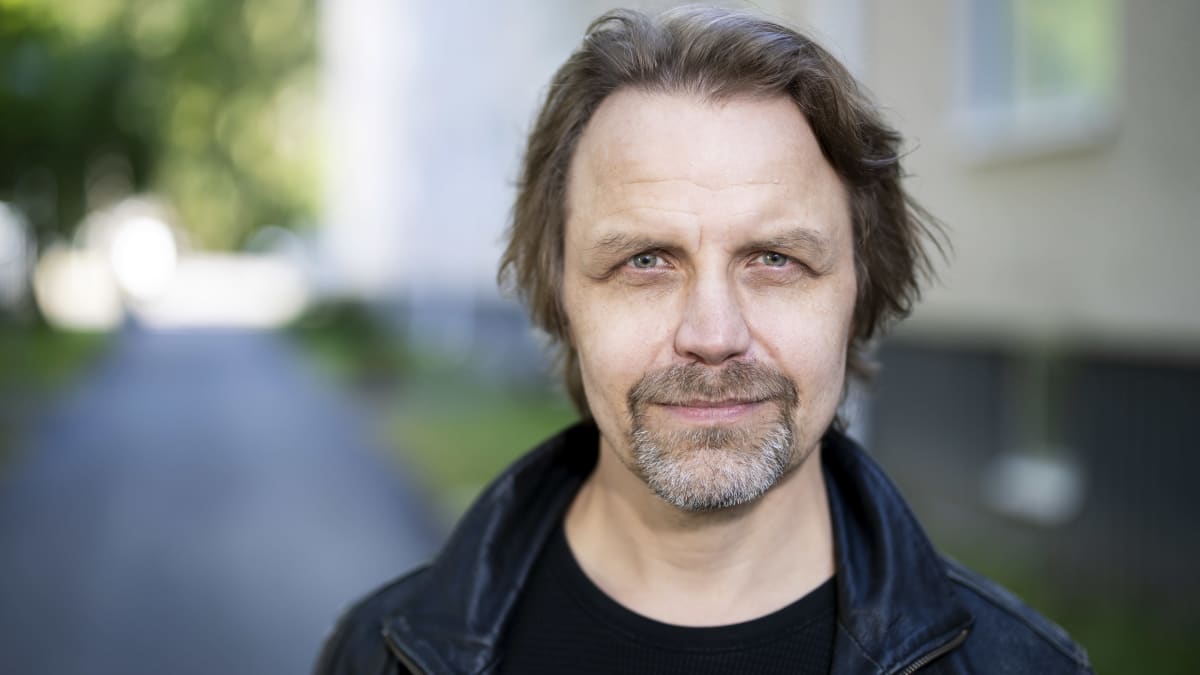 Mies katsoo suoraan kameraan kerrostalon pihalla. 