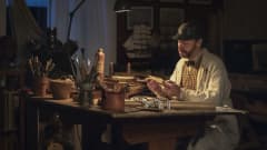 Näyttelijä Peter Franzén pöydän ääressä tutkimassa lappua. Pöydällä kynäpurkkeja, paperirulla. 