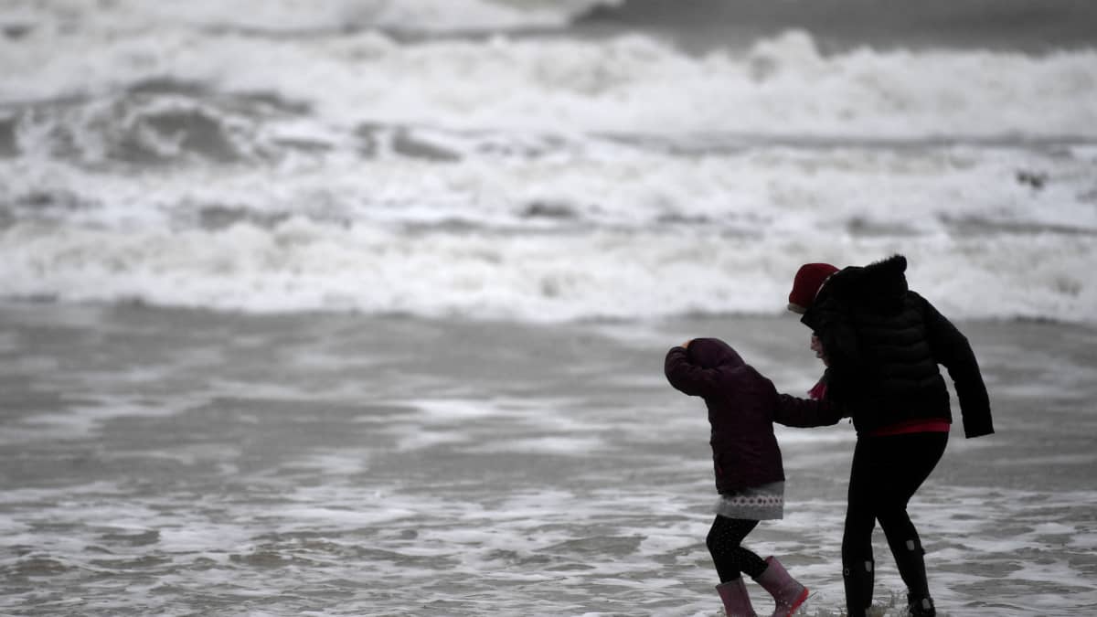 Aikuinen ja lapsi kävelevät kovassa tuulessa rannalla. Taustalla myrskyävä meri.
