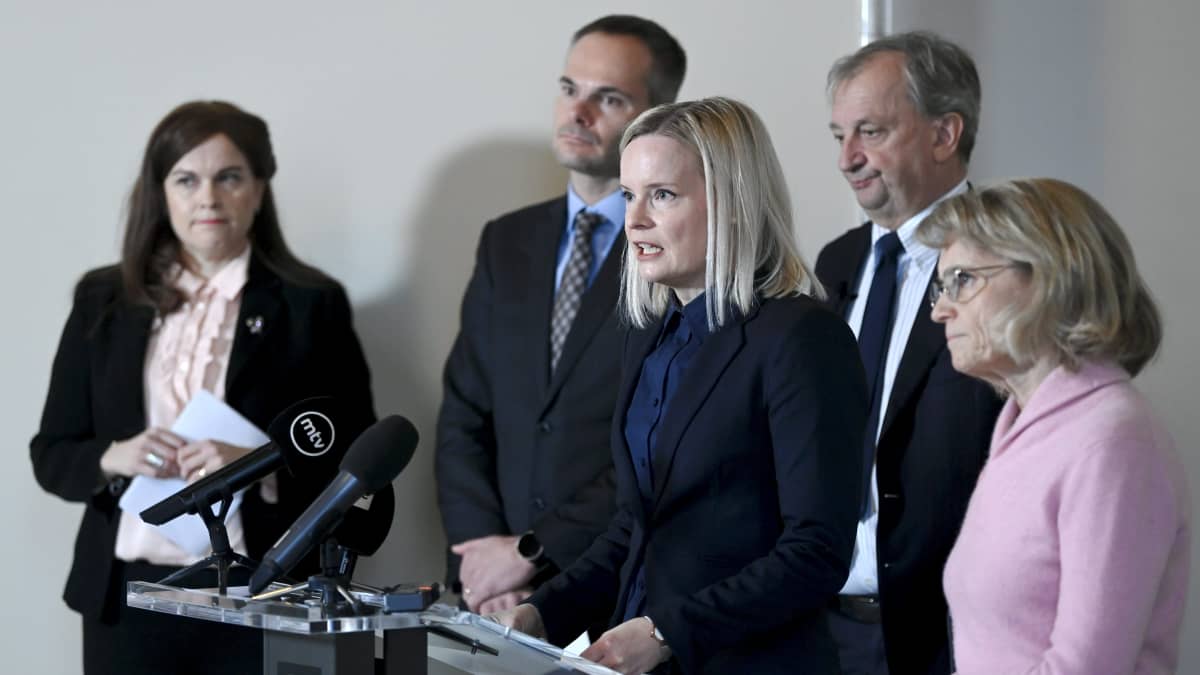 Riikka Purra puhuu mikrofoniin, ympärillä opposition edustajia.