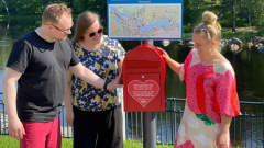 Mies ja kaksi naista katsovat punaista postilaatikkoa kesäisen joen rannalla.