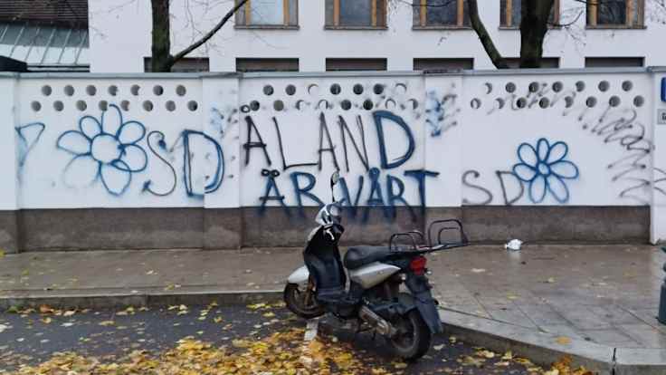 A pale grey wall with round perforations and graffiti reading "Åland är vårt" and "SD" with flowers. In the foreground, a parked motorcycle and yellow leaves on the street.