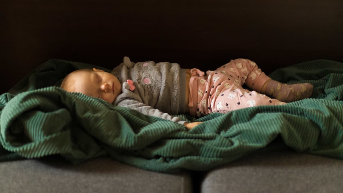 Harmaaseen puseroon ja pinkkeihin housuihin puettu vauva nukkuu vihreällä peitolla.