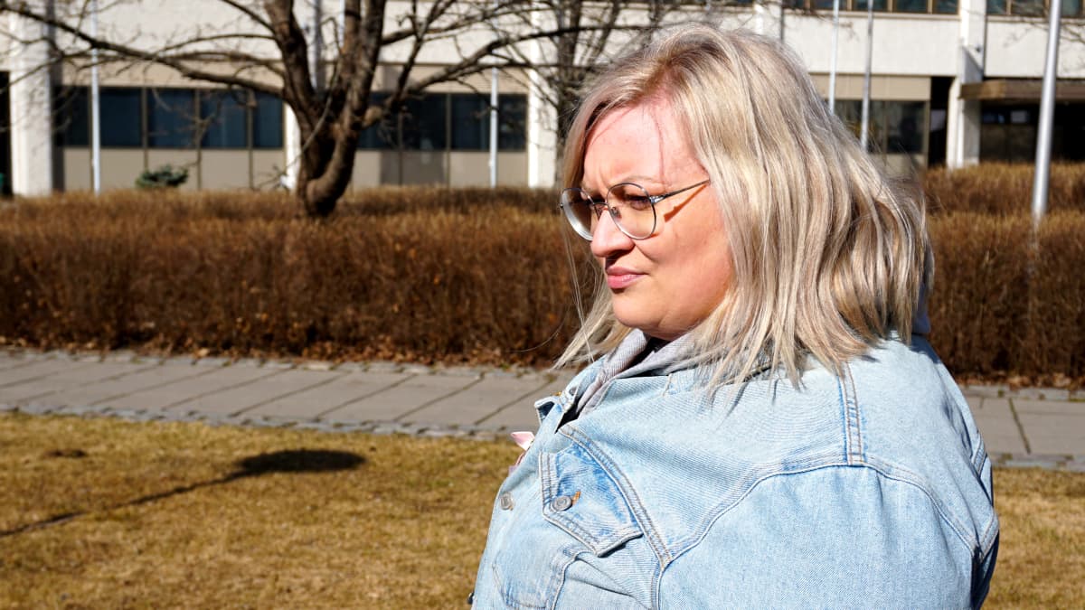 Blond kvinna i glasögon och jeansjacka står i en park.