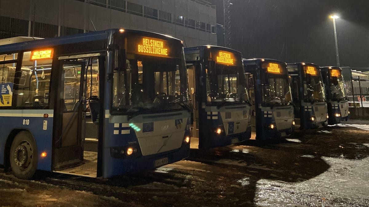 Viisi Ukrainaan lähtevää sinistä avustusbussia seisoo Länsisatamassa odottamassa lastausta. Pimeä, luminen ilma taustalla.