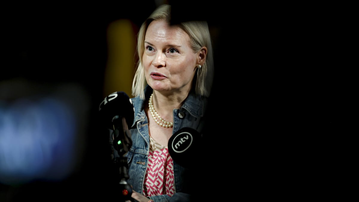 Riikka Purra wearing a denim jacket, red-and-white shirt and pearl necklace against a dark background while speaking to reporters at the House of Estates on 24 May 2023.