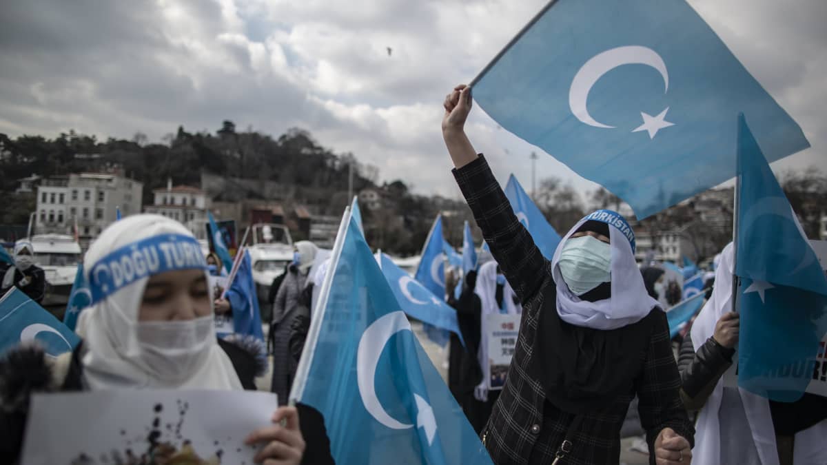 En kvinna med huvudduk och munskydd viftar med uigurernas blåa flagga.