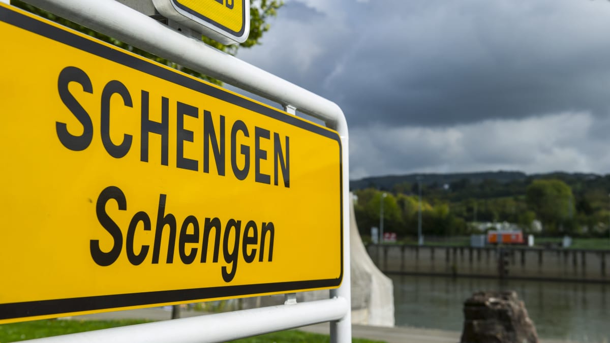 Vasemmalla etualalla isona keltainen Luxemburgissa kuvattu Scengen-kyltti, jonka oikealla puolella sumeaa maisemaa.