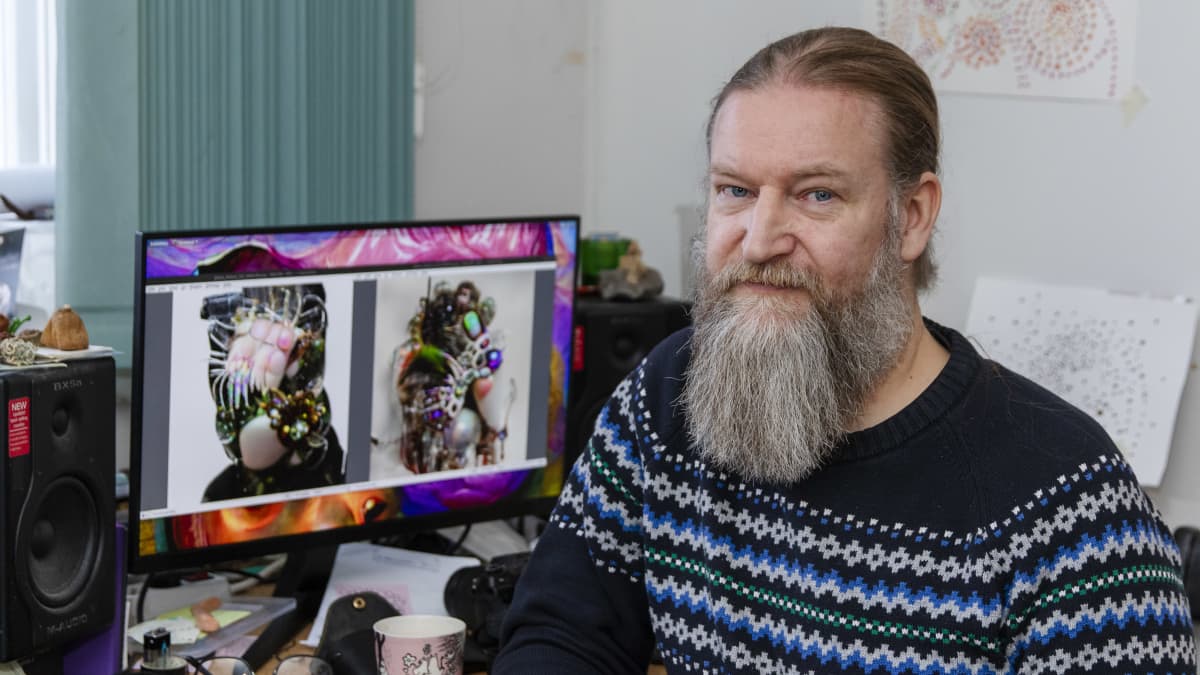 Parrakas Jukka Hautamäki istuu työpöytänsä ääressä. Taustalla olevassa tietokoneen monitorissa näkyy kaksi hänen digitaalista taideteostaan.