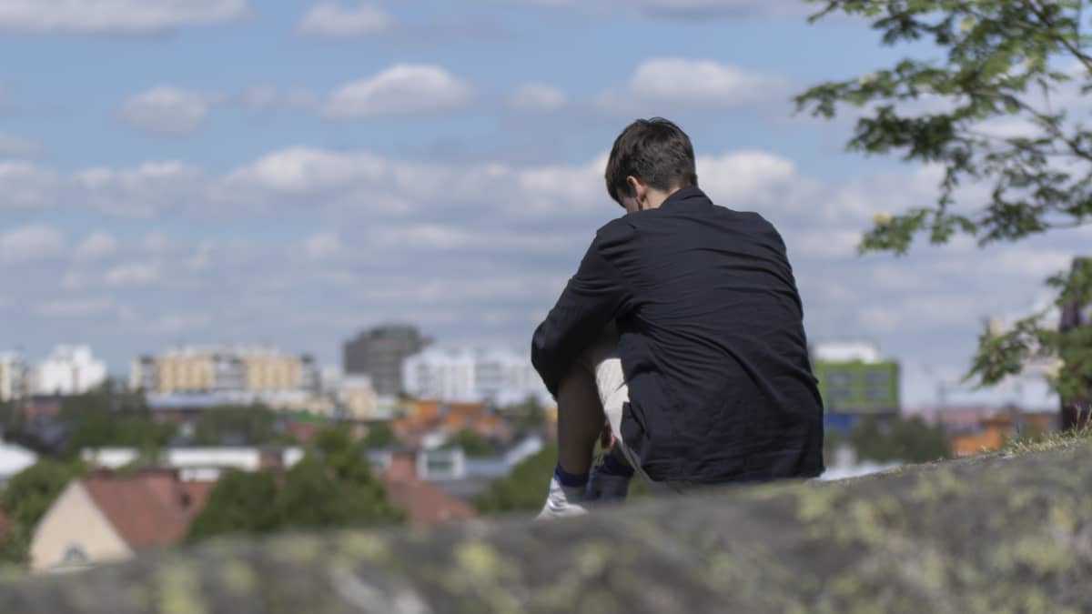 Nuori mies istuu kalliolla. Taustalla siintää taloja ja taivas.