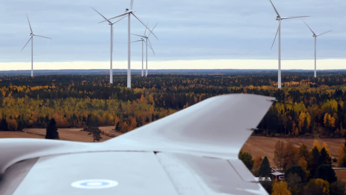Ilmavoimien Pilatus PC-12 NG -yhteyskoneen siipi, jonka taustalla näkyy tuulivoimaloita syksyisessä maaseudun metsämaisemassa.