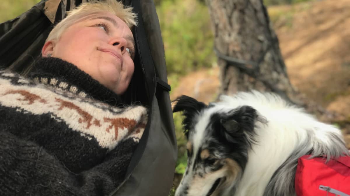 Riippumatossa makaava nainen hymyilee koiralle, joka istuu vieressä