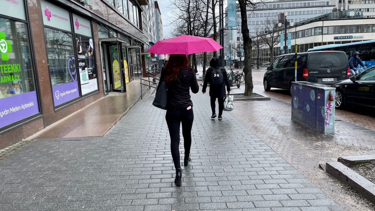 Nuori nainen pitää aniliininpunaista satenvarjoa ja kävelee kadulla poispäin kamerasta