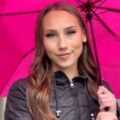 Nuori nainen, jolla n pitkät ruskeat hiukset ja upeasti meikatut silmät katsoo kameraan. Pään päällä on auki aniliininpunainen sateenvarjo. 