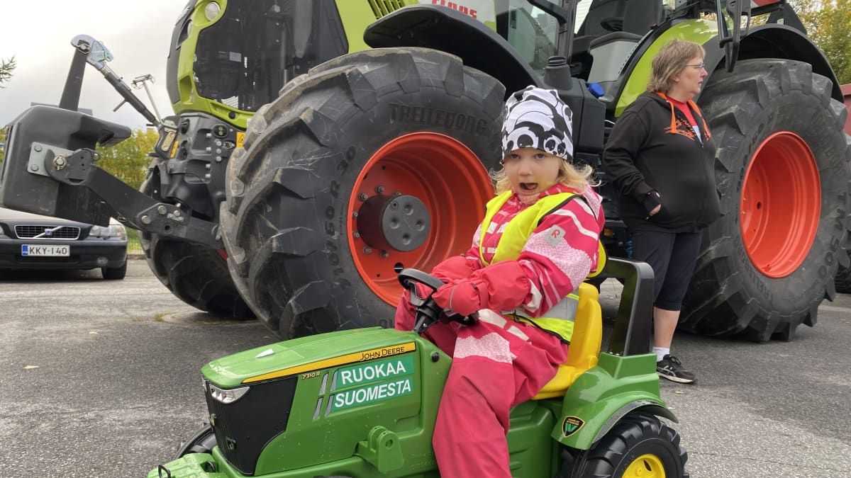 Pieni tyttö, Enni Herajärvi, istuu ulkoiluhaalarissaan ohjaamassa polkutraktoria. Taustalla oikea traktori ja MTK-Tornion puheenjohtaja Anne-Mari Söderström.